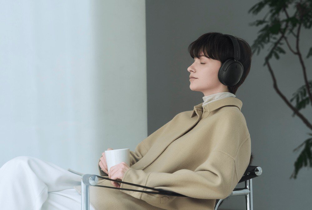 Słuchawki nauszne SONY WH-CH735N Kobieta w beżowej kurtce siedzi na przezroczystym krześle przy oknie. Trzyma biały kubek, ma zamknięte oczy i czarne słuchawki nagłowne. W tle widać roślinę