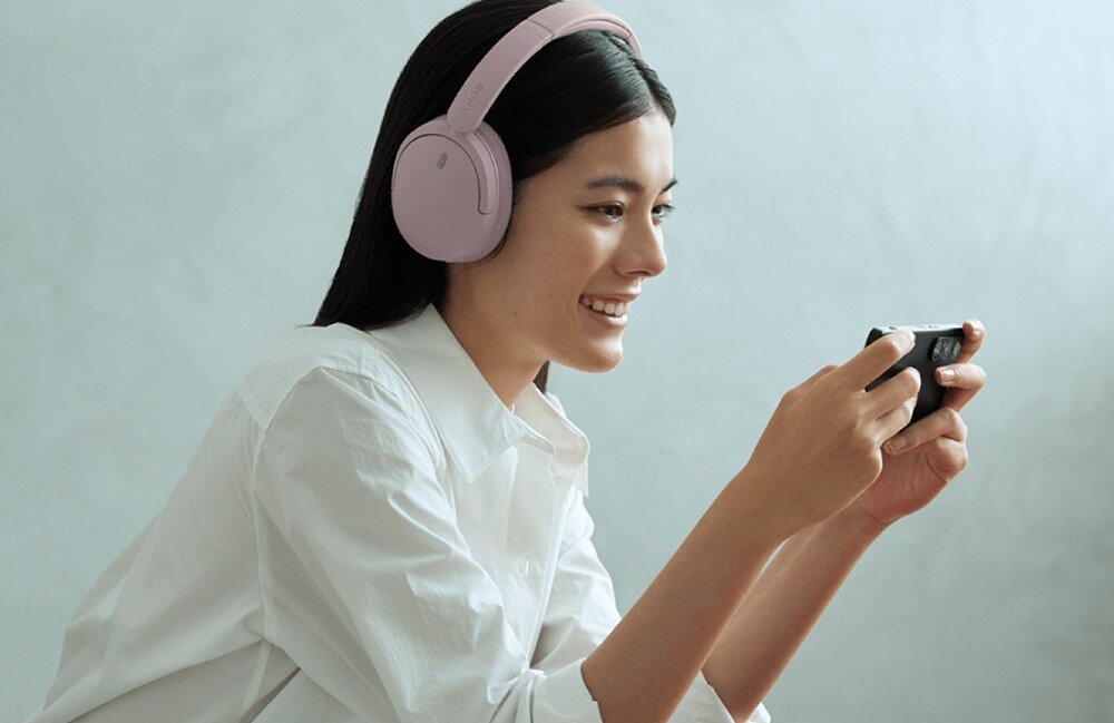 Słuchawki nauszne SONY WH-CH735N Kobieta w białej koszuli siedzi bokiem, patrząc w bok. Trzyma smartfon oburącz, na uszach ma jasnoróżowe słuchawki nagłowne. Tło jest jasne i gładkie