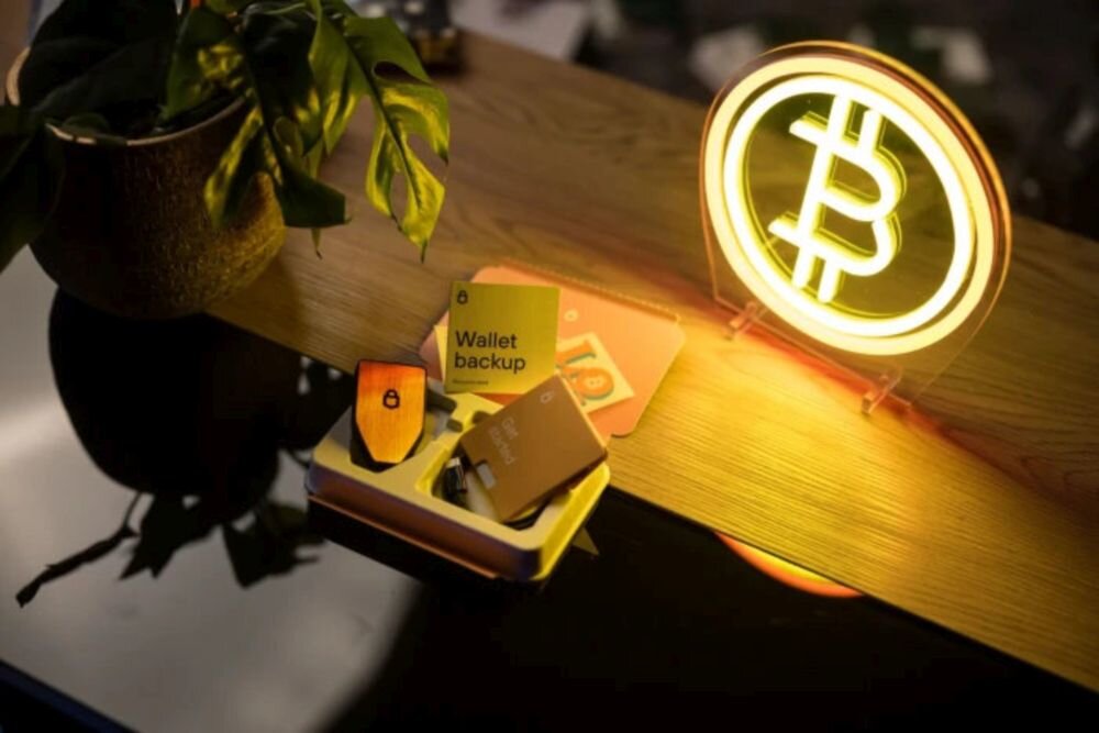 Na drewnianym blacie znajduje się otwarte pudełko z urządzeniem elektronicznym oraz karta z napisem \'Wallet backup\'. Obok stoi świecący symbol bitcoina, a w tle widoczna jest roślina w doniczce.