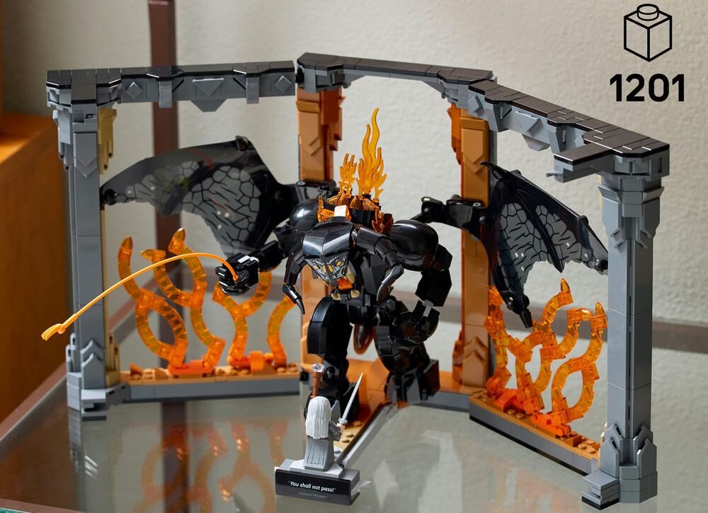 Zestaw LEGO przedstawiający scenę z dużą, czarną postacią z płomieniami i skrzydłami oraz mniejszą, białą figurką z mieczem. W tle widoczne są pomarańczowe płomienie i szara konstrukcja, a na podstawce napis \'You shall not pass!\'.