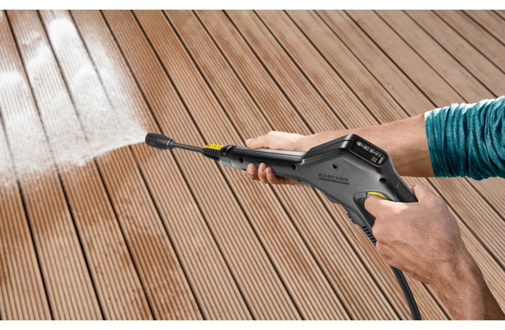 Osoba trzymająca myjkę ciśnieniową, z której wydobywa się strumień wody, skierowany na drewnianą powierzchnię. Na myjce widoczne jest logo \'KARCHER\'.