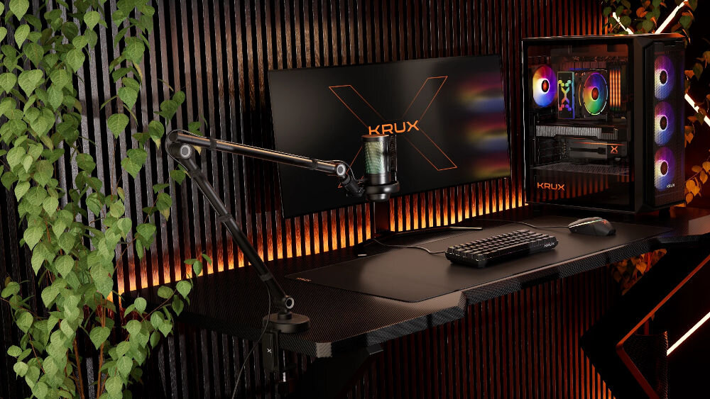 Na biurku znajduje się mikrofon na wysięgniku, monitor z logo \'KRUX\' oraz komputer z podświetleniem RGB. W tle widoczne są rośliny i ściana z pionowymi panelami.
