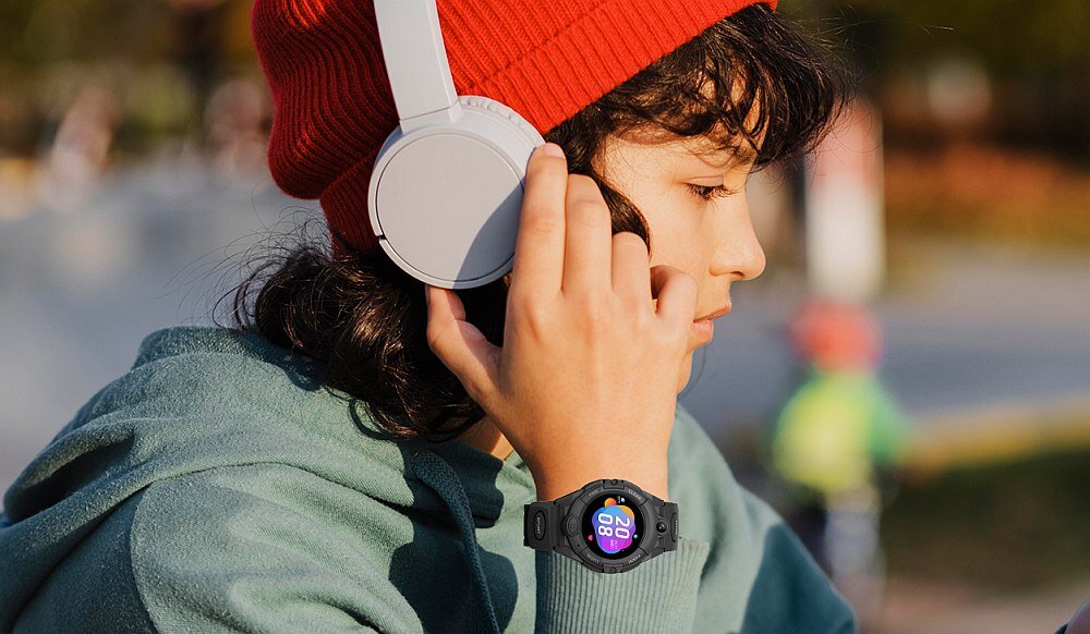 Osoba nosi słuchawki i smartwatch z kolorowym wyświetlaczem, na którym widoczna jest godzina 20:08. W tle rozmyty krajobraz na zewnątrz.