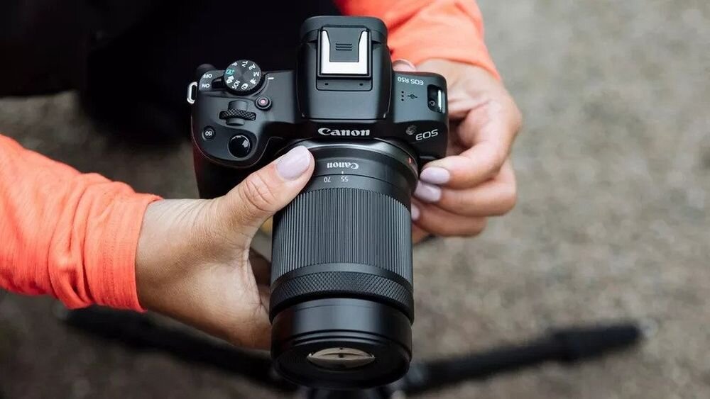 Osoba trzyma aparat bezlusterkowy Canon EOS z zamontowanym obiektywem. Na aparacie widoczne są napisy: \'Canon\' i \'EOS\'.