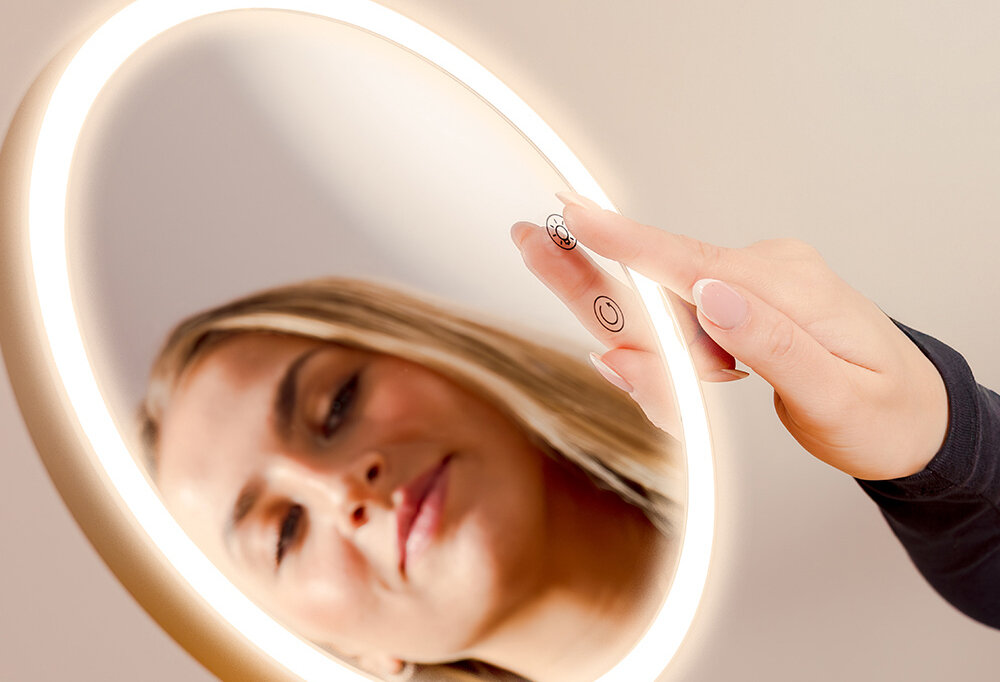 Osoba dotyka przycisku na lusterku kosmetycznym z podświetleniem LED. Na lusterku widoczne są dwa symbole: okrąg z kreską i okrąg z trzema liniami.
