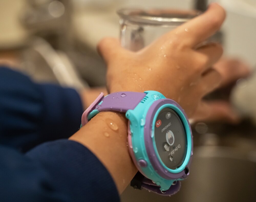 Osoba nosi niebieski smartwatch z fioletowym paskiem, na którym widoczny jest ekran z ikonami. Na ręce są krople wody, a w tle znajduje się szklanka.