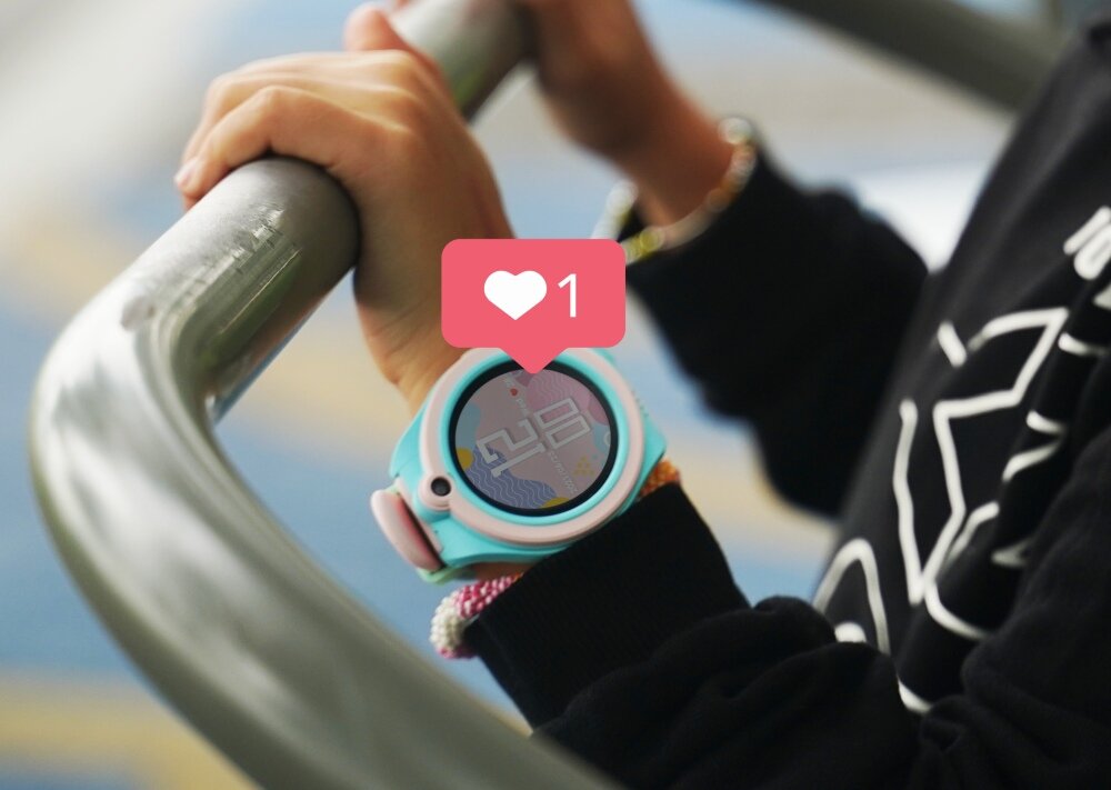 Osoba trzyma metalową poręcz, na nadgarstku ma niebieski smartwatch z wyświetlaczem pokazującym godzinę. Na ekranie zegarka widoczna jest ikona serca z liczbą \'1\'.