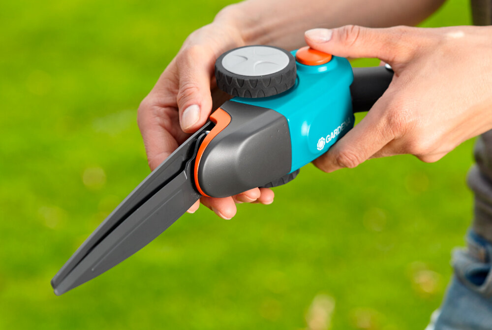 Osoba trzyma ręczne nożyce do trawy w kolorze niebiesko-czarnym z pomarańczowym przyciskiem. Na narzędziu widoczne jest logo \'GARDENA\'.