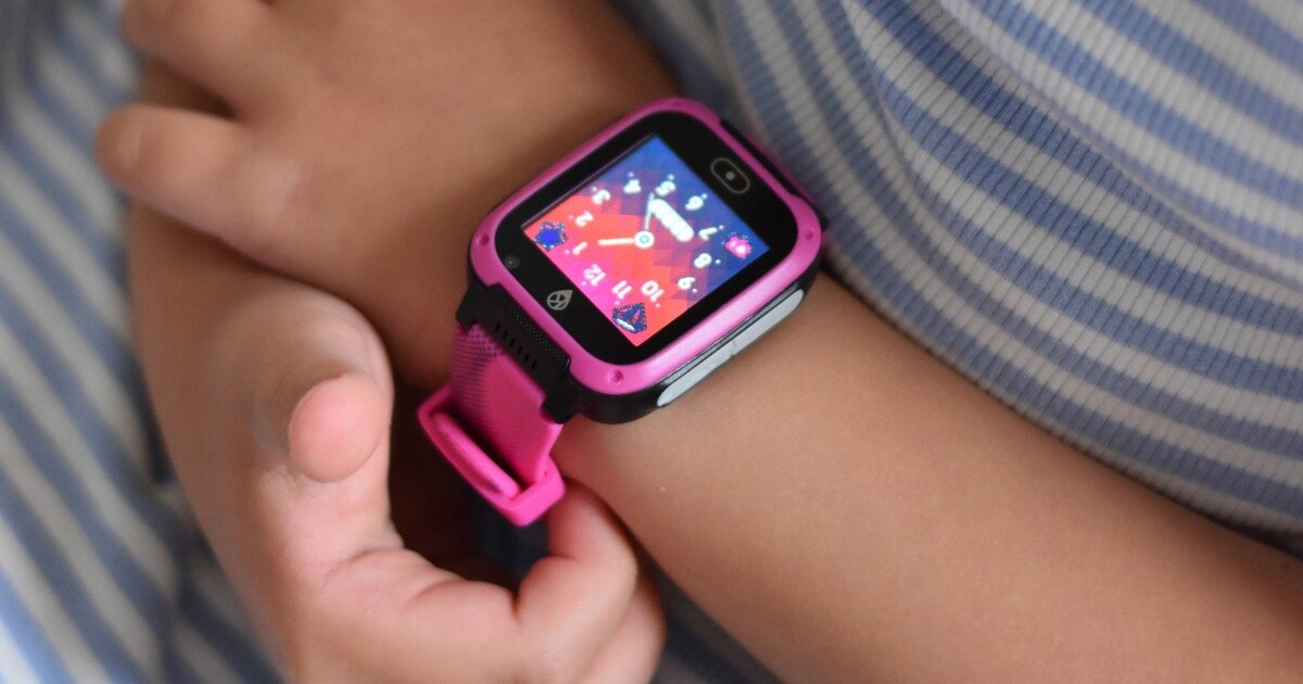 Na zdjęciu widoczny jest różowy smartwatch na nadgarstku dziecka, z kolorowym wyświetlaczem pokazującym tarczę zegara. Pasek zegarka jest również różowy, a tło stanowi materiał w paski.