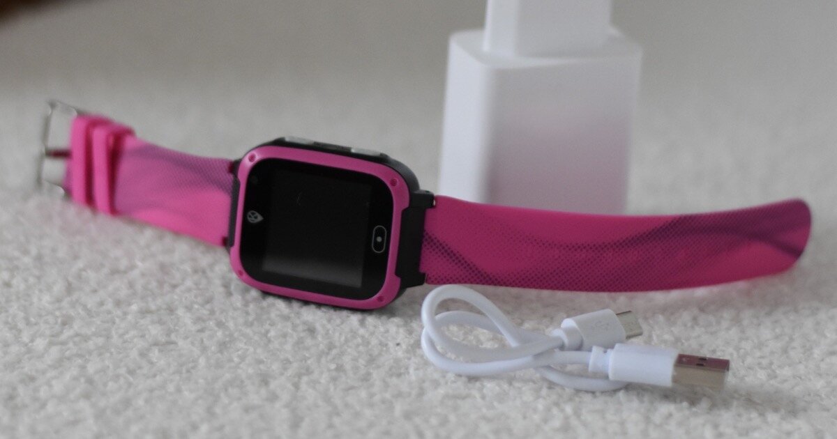Różowy smartwatch z czarnym ekranem leży na jasnej powierzchni obok białego kabla USB i ładowarki.