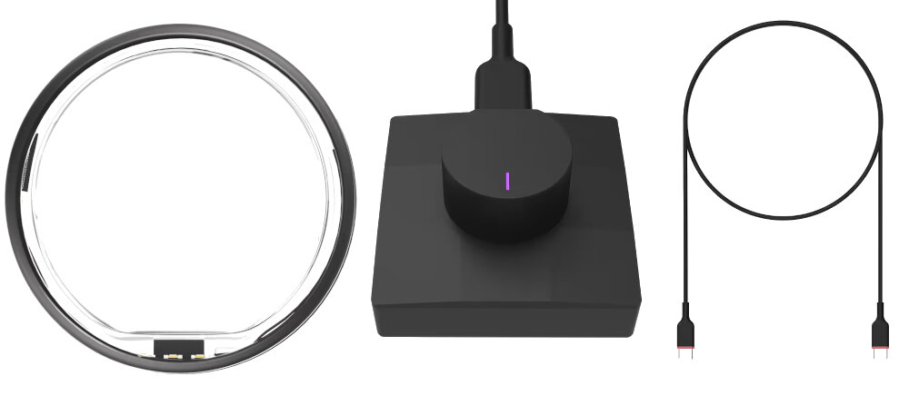 Obraz przedstawia matowo-szary pierścień, czarną stację ładującą z podłączonym kablem oraz osobny kabel z końcówkami USB. Stacja ładująca ma okrągły element z fioletowym wskaźnikiem.