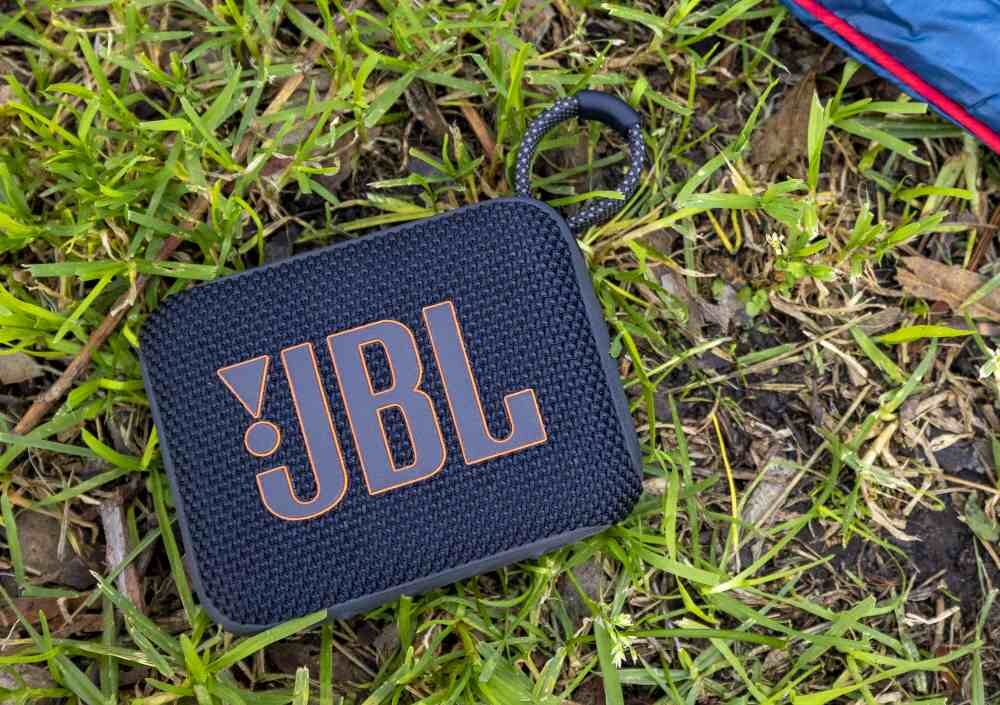 Czarny głośnik przenośny z logo \'JBL\' leży na trawie. Głośnik ma teksturowaną powierzchnię i przypięty sznurek.