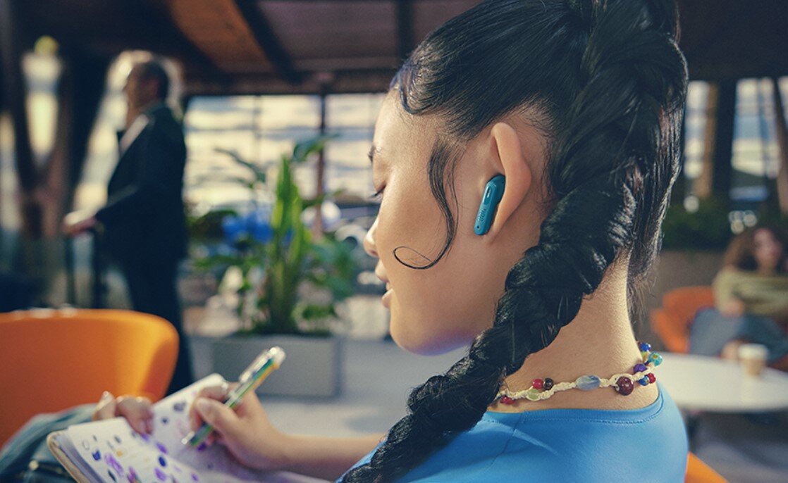 Osoba z długim warkoczem nosi czarne słuchawki dokanałowe. W tle widać rośliny i osoby siedzące na pomarańczowych fotelach.