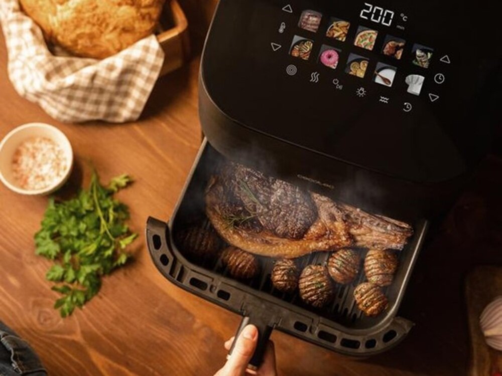 Na drewnianym stole znajduje się otwarta frytkownica beztłuszczowa z wyświetlaczem pokazującym temperaturę 200°C oraz różne ikony. W środku frytkownicy widać kawałek mięsa i ziemniaki, a obok leży natka pietruszki i miseczka z przyprawami.