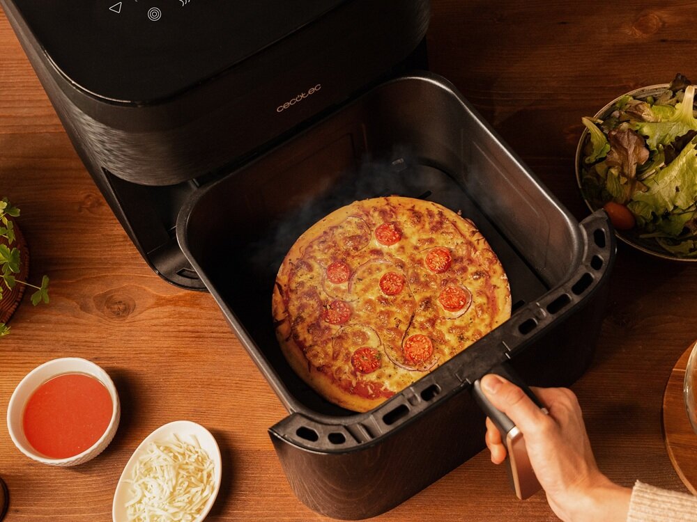 Na drewnianym blacie stoi frytkownica beztłuszczowa z otwartą szufladą, w której znajduje się pizza z pomidorami. Obok widoczne są miski z sosem pomidorowym, startym serem i sałatką.