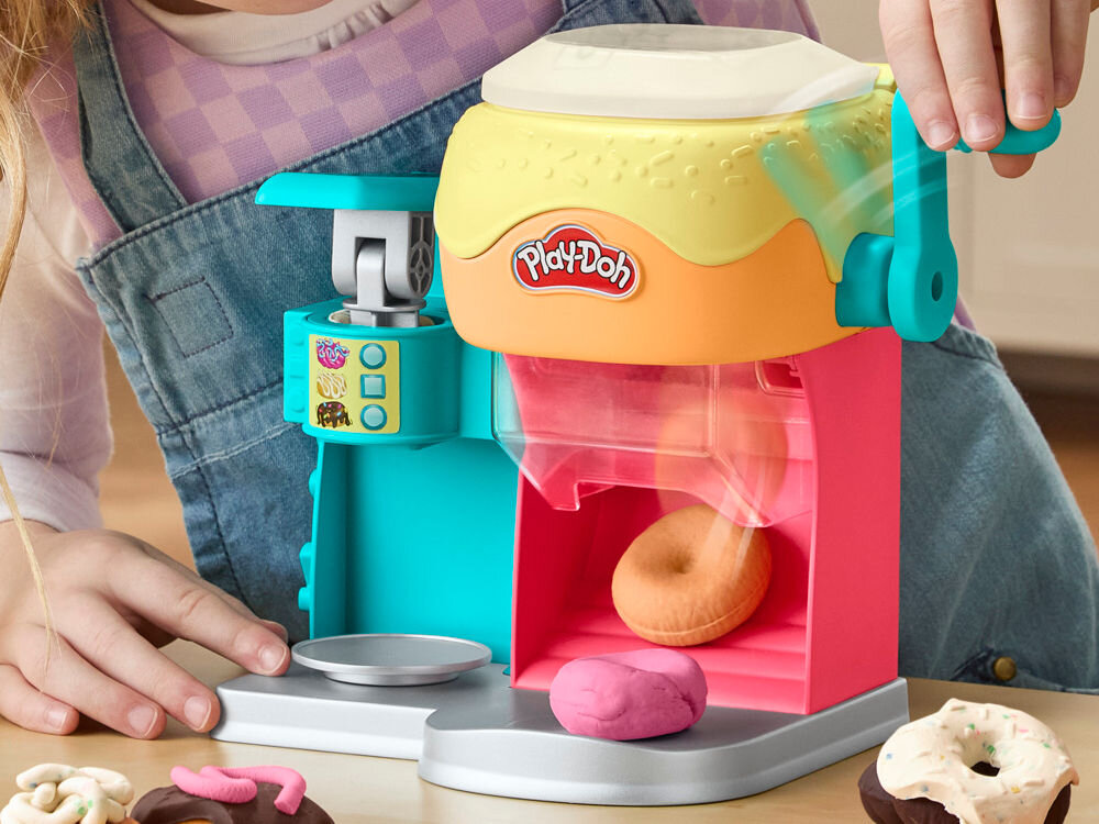 Zabawka do tworzenia pączków z ciastoliny z logo \'Play-Doh\' na górze. Widać ręce dziecka obsługujące urządzenie, a na tacy leżą kolorowe pączki z ciastoliny.