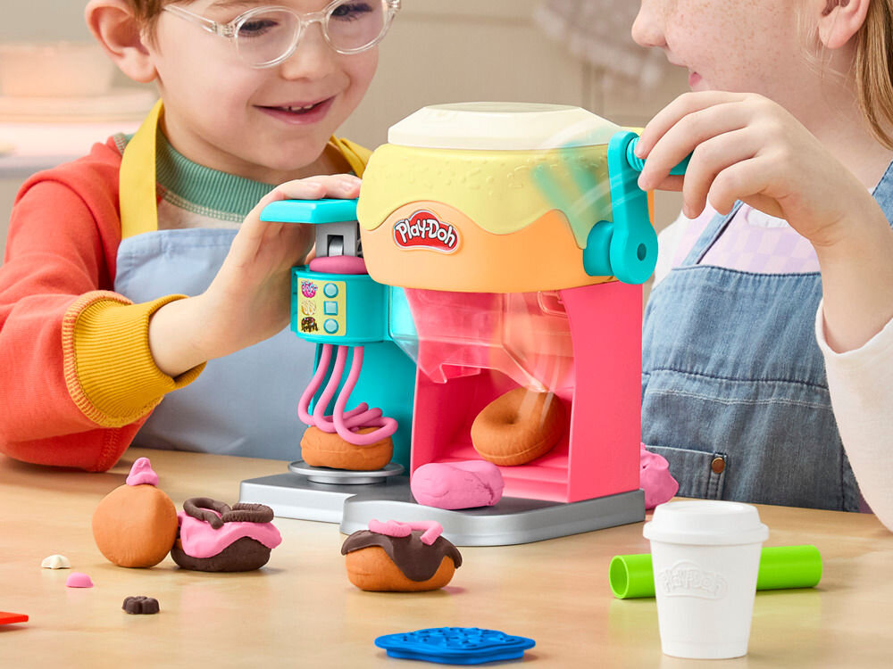 Dwoje dzieci bawi się zestawem do tworzenia pączków z ciastoliny Play-Doh, który ma kolorową maszynę z logo Play-Doh. Na stole znajdują się różne elementy z ciastoliny, w tym pączki i kubek.