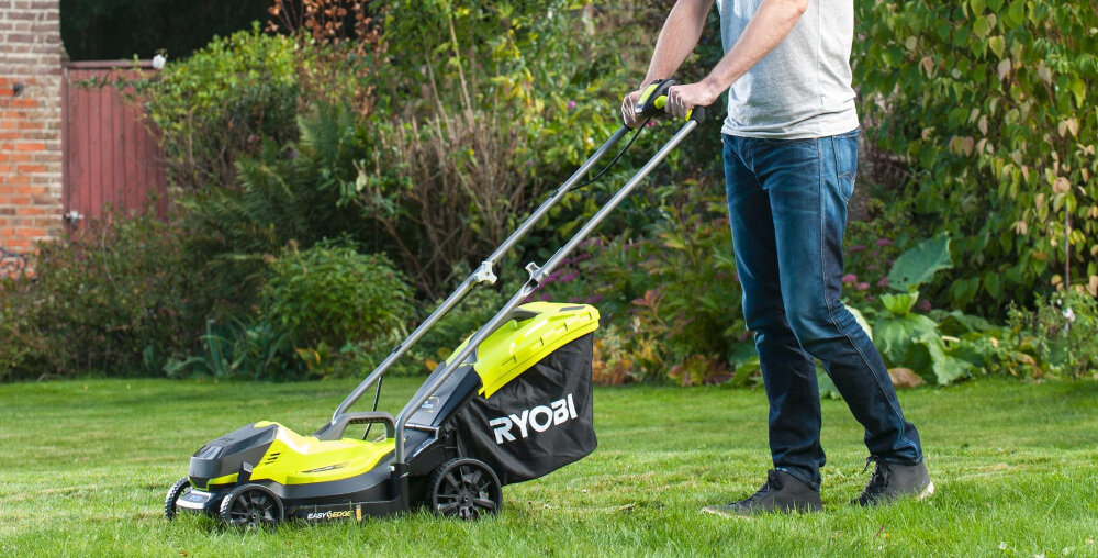 Osoba pcha kosiarkę akumulatorową z widocznym logo \'RYOBI\' na trawie w ogrodzie. Kosiarka ma czarno-zielony kolor i duże koła.