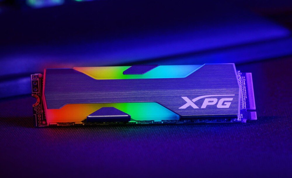 Na obrazie widoczny jest dysk SSD z podświetleniem RGB, z logo \'XPG\' na obudowie. Dysk leży na ciemnym tle, a jego podświetlenie emituje różnokolorowe światło.