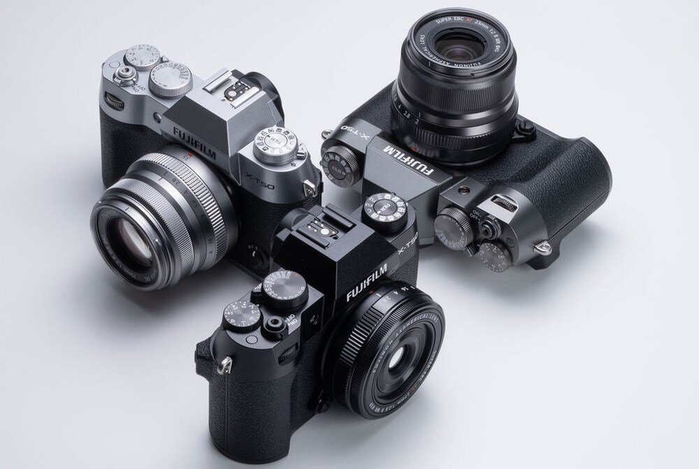 Na obrazie widoczne są trzy aparaty fotograficzne marki Fujifilm, każdy z obiektywem. Aparaty mają różne kolory i są ustawione na jasnym tle.