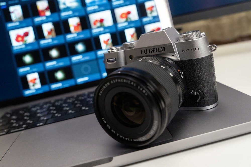 Na zdjęciu widać aparat bezlusterkowy z obiektywem, leżący na otwartym laptopie. Na korpusie aparatu widoczne są napisy \'FUJIFILM\' i \'X-T50\'.