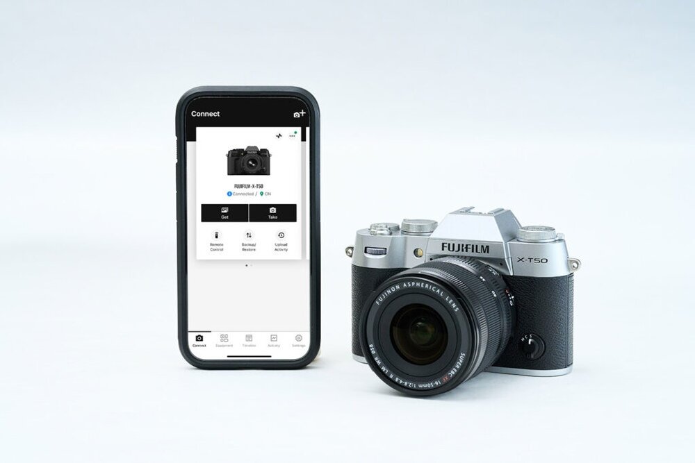 Na obrazie widoczny jest czarny aparat bezlusterkowy z obiektywem oraz smartfon z aplikacją pokazującą ten sam model aparatu. Na obiektywie widoczny jest napis \'FUJINON ASPHERICAL LENS\'.