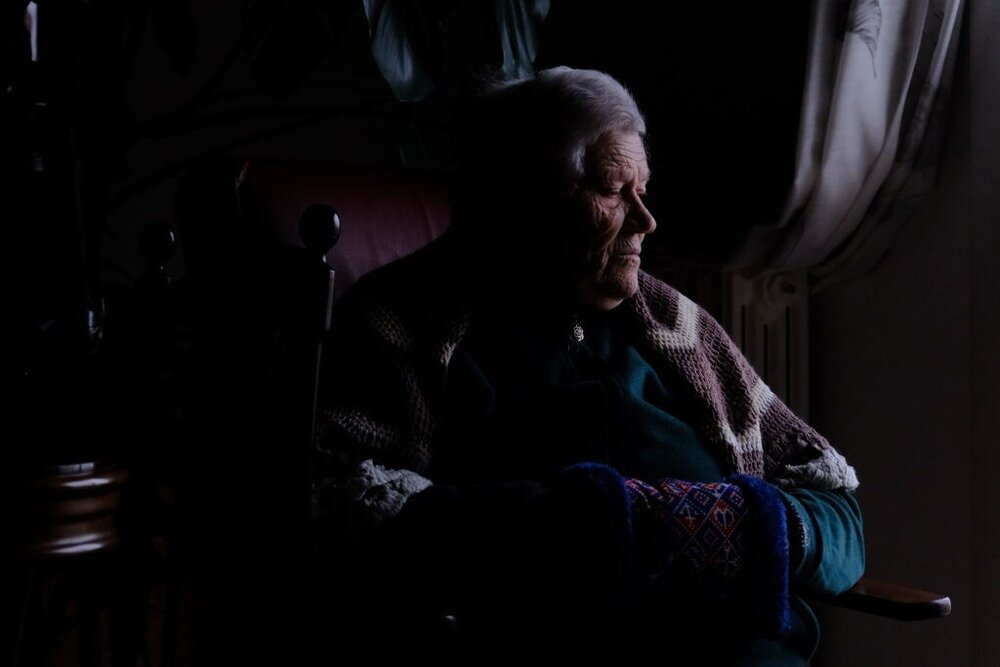 Osoba siedzi w fotelu, patrząc w stronę okna, przez które wpada światło. Na ramionach ma narzucony koc, a ręce są schowane w rękawicach.