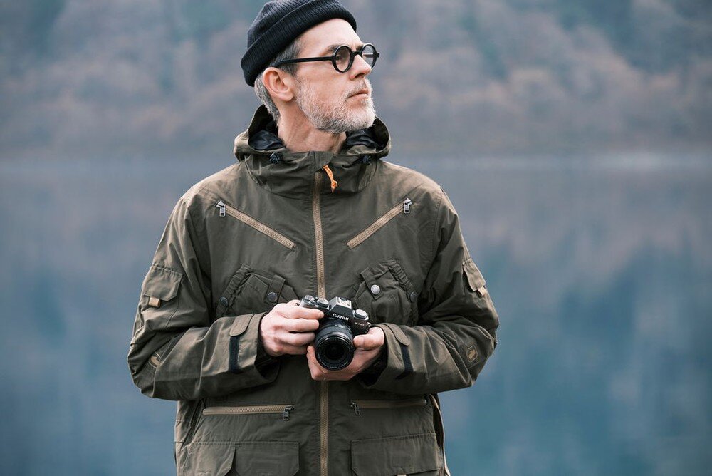 Osoba w ciemnej kurtce i czapce trzyma aparat fotograficzny. W tle widać rozmyty krajobraz z wodą i drzewami.