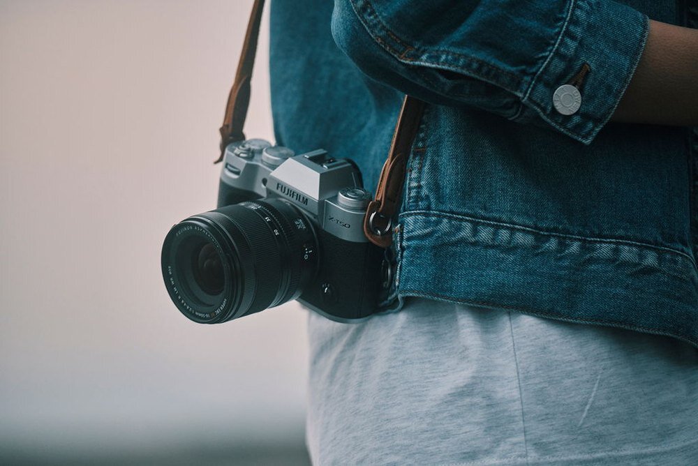 Osoba nosi aparat fotograficzny z obiektywem, zawieszony na pasku. Aparat ma widoczne logo \'Fujifilm\' i jest przymocowany do brązowego paska, a osoba ma na sobie niebieską kurtkę jeansową.