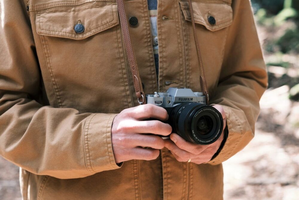Osoba w brązowej kurtce trzyma aparat fotograficzny z widocznym logo \'FUJIFILM\'.