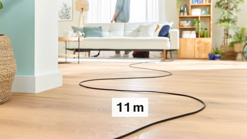 Na drewnianej podłodze widać długi czarny kabel z oznaczeniem \'11 m\'. W tle znajduje się jasna sofa, a obok niej rośliny i regał z książkami.