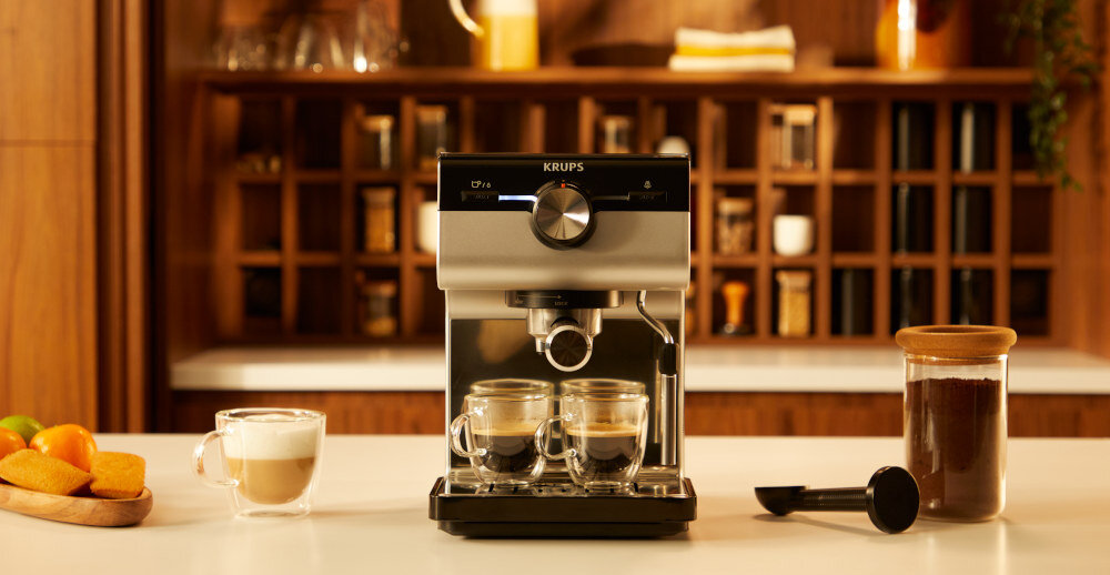 Na blacie kuchennym stoi ekspres do kawy marki KRUPS z dwoma filiżankami espresso. Obok znajduje się szklanka z kawą mleczną, słoik z kawą mieloną i łyżka do kawy.