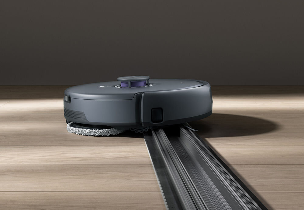 Na drewnianej podłodze znajduje się ciemnoszary robot sprzątający z okrągłym kształtem, wyposażony w szczotki czyszczące. Robot porusza się po metalowej szynie.