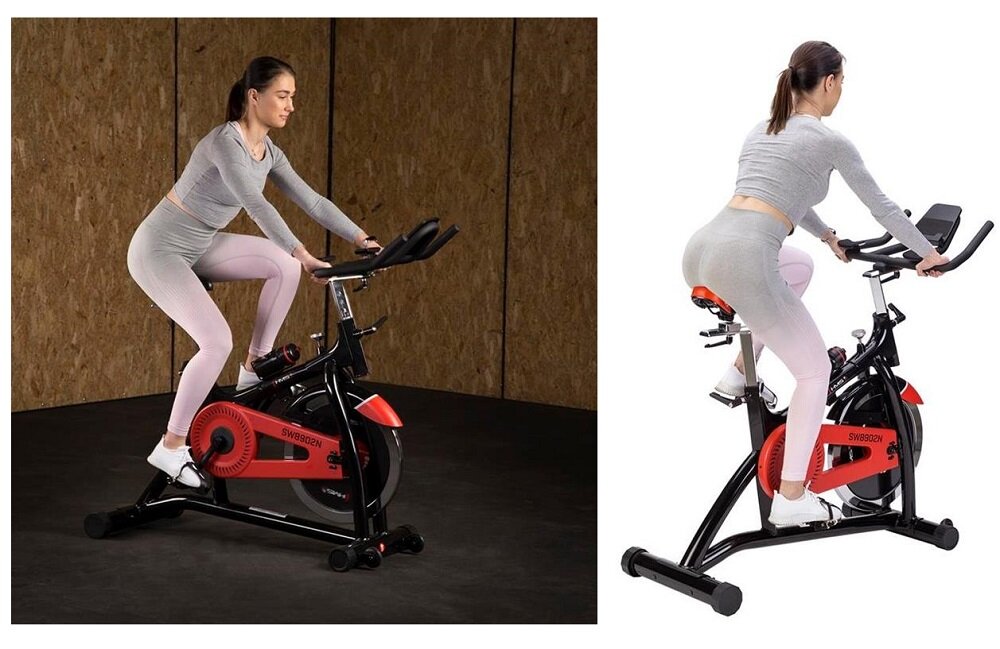 Dwie kobiety ćwiczą na rowerze spinningowym w pomieszczeniu. Rower ma czarną ramę z czerwonymi elementami i widoczne logo \'SW8902N\'.