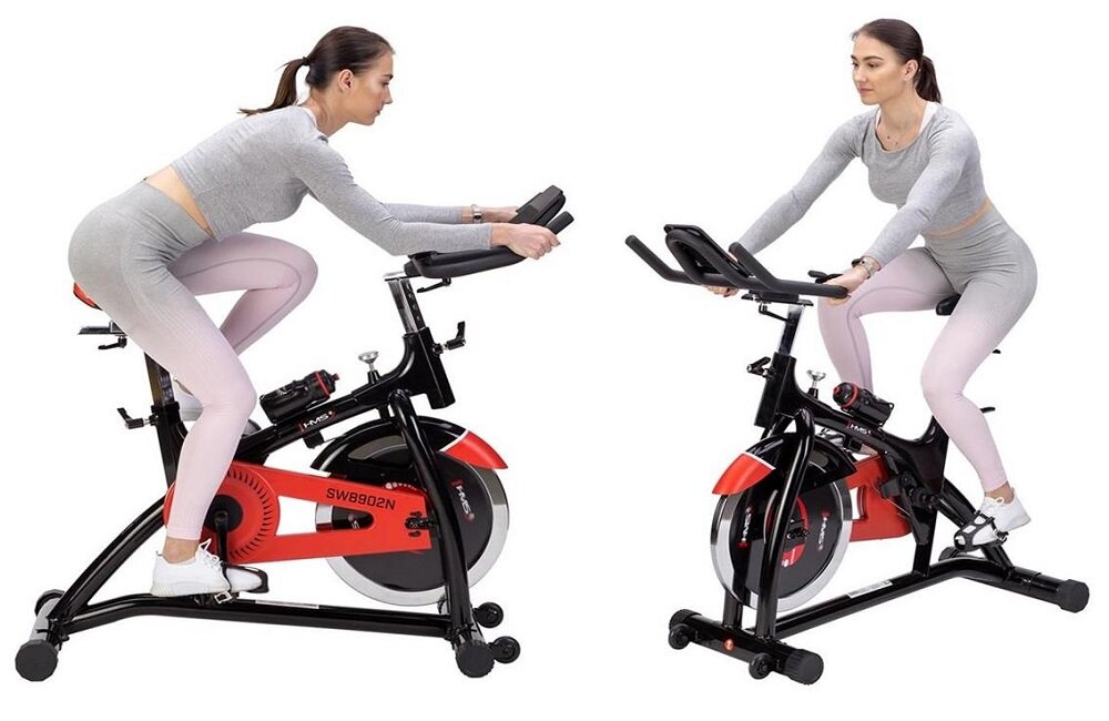 Dwie kobiety ćwiczą na rowerach spinningowych HMS SW8902N w kolorze czarno-czerwonym. Na ramie roweru widoczny jest napis \'SW8902N\'.
