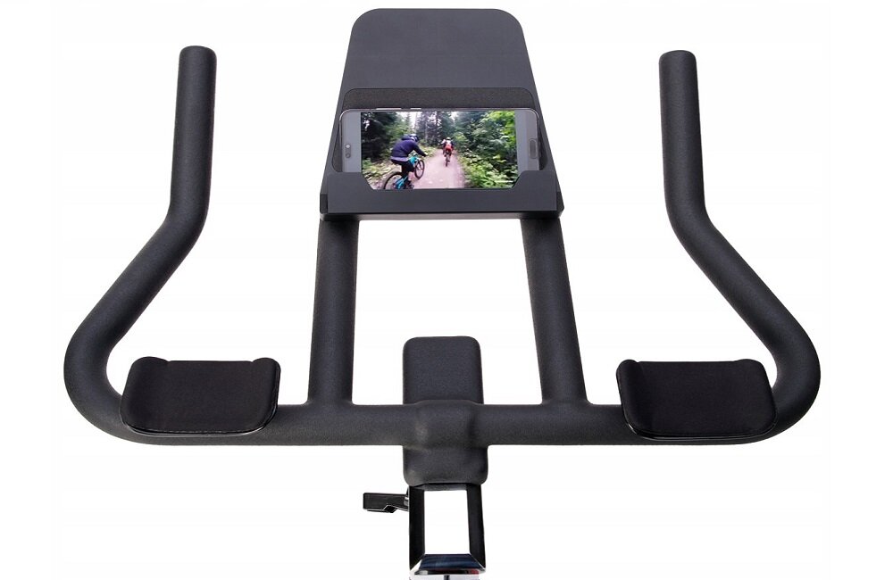 Kierownica roweru spinningowego z uchwytem na urządzenie mobilne, na którym wyświetlany jest obraz z jazdy rowerowej w lesie. Kierownica ma dwa uchwyty i podkładki na dłonie.