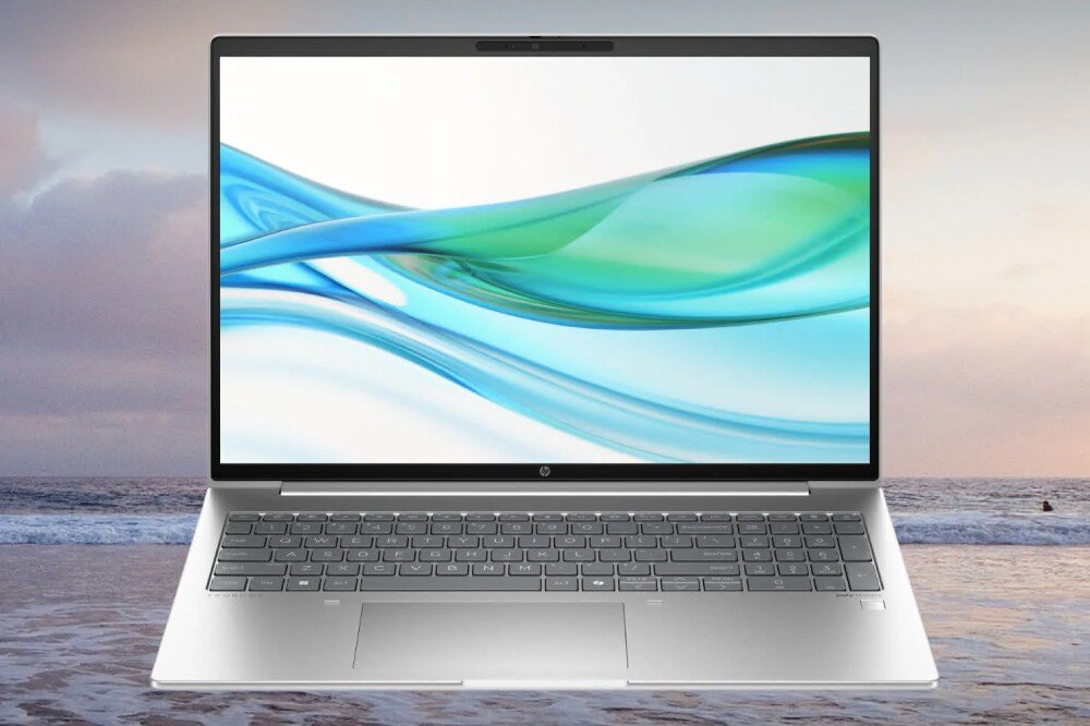 Laptop z dużym ekranem wyświetlającym abstrakcyjny wzór w odcieniach niebieskiego i zielonego. W tle widoczny jest krajobraz plaży z morzem i niebem.