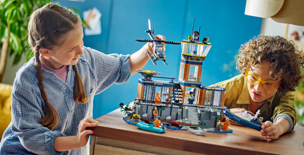 Dwoje dzieci bawi się zestawem klocków LEGO przedstawiającym więzienną wyspę z wieżą strażniczą i helikopterem. W tle widać niebieską ścianę i roślinność.