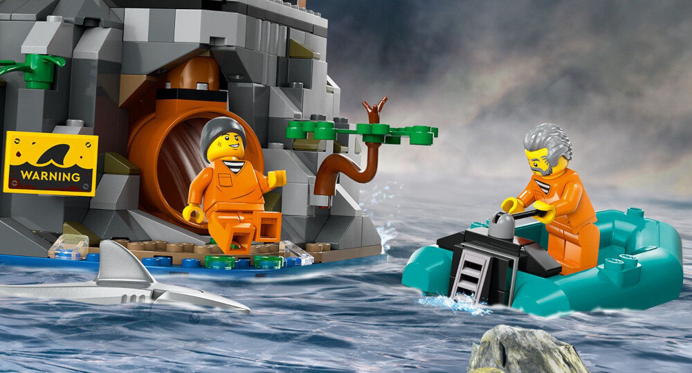 Na obrazie widać zestaw klocków LEGO przedstawiający scenę z więziennej wyspy, z dwoma figurkami w pomarańczowych strojach i rekinem w wodzie. Na skale znajduje się żółty znak z napisem \'WARNING\'.