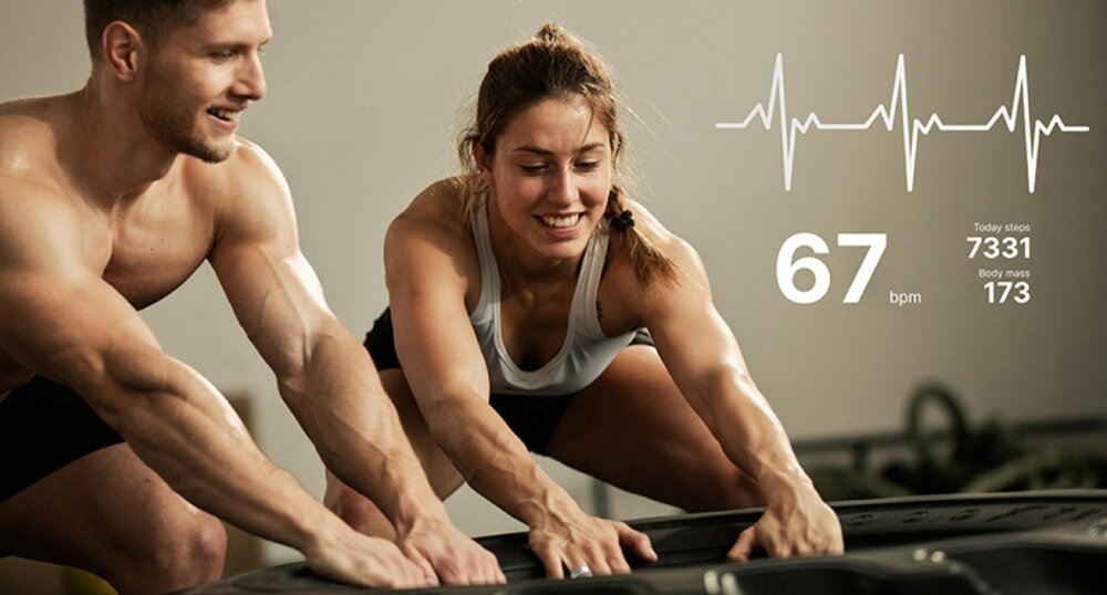 Dwie osoby ćwiczą, pchając duży obiekt. Na obrazie widoczny jest wykres tętna oraz tekst: \'67 bpm\', \'Today steps 7331\', \'Body mass 173\'.