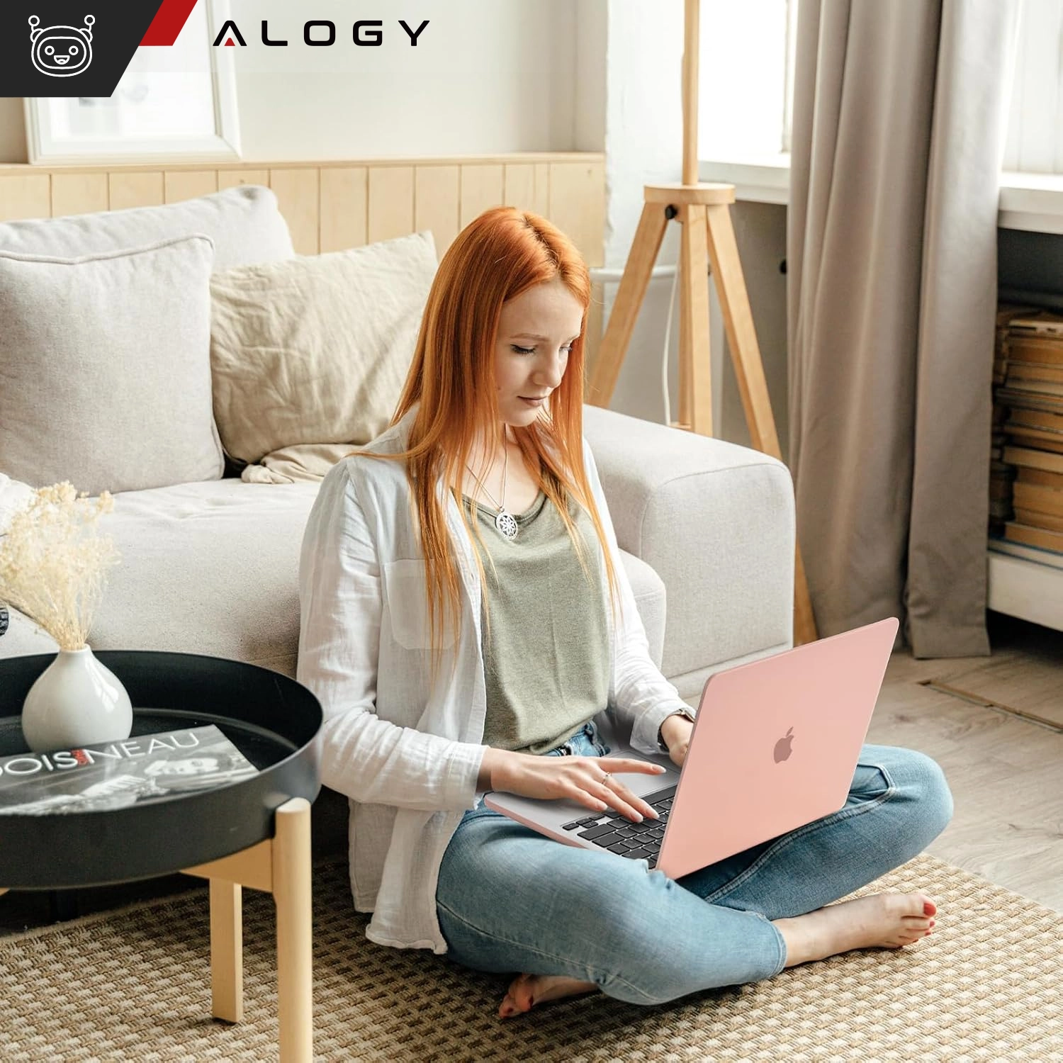 Osoba siedzi na podłodze w salonie, używając laptopa z różowym, przezroczystym etui. W tle widoczna jest sofa, stolik z czasopismem i wazonem oraz logo \'ALOGY\'.