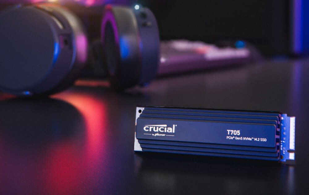 Na ciemnym tle znajduje się dysk SSD z radiatorem z napisem \'crucial by micron T705 PCIe Gen5 NVMe M.2 SSD\'. W tle widoczne są słuchawki i rozmyte światła.