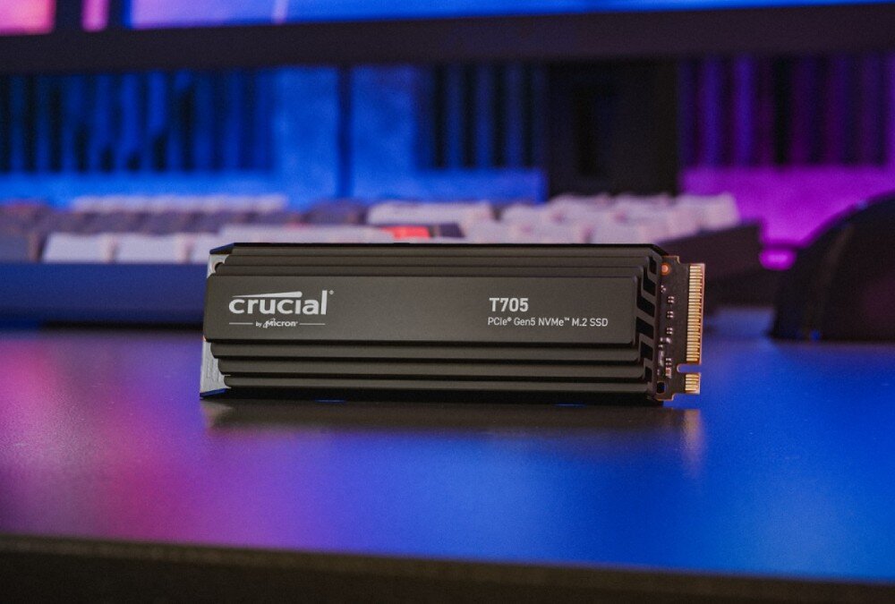 Na zdjęciu znajduje się dysk SSD z radiatorem, na którym widnieje napis \'crucial by micron T705 PCIe Gen5 NVMe M.2 SSD\'. W tle widać rozmytą klawiaturę i kolorowe oświetlenie.