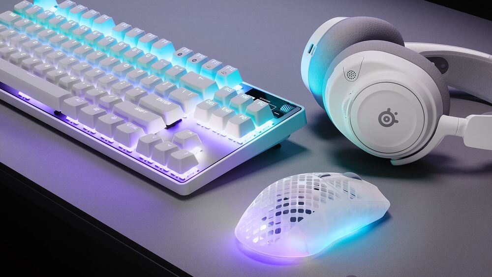 Na obrazie znajduje się klawiatura z podświetleniem RGB, mysz z ażurową obudową i podświetleniem oraz słuchawki z logo SteelSeries. Wszystkie urządzenia są w jasnej kolorystyce i leżą na ciemnym blacie.