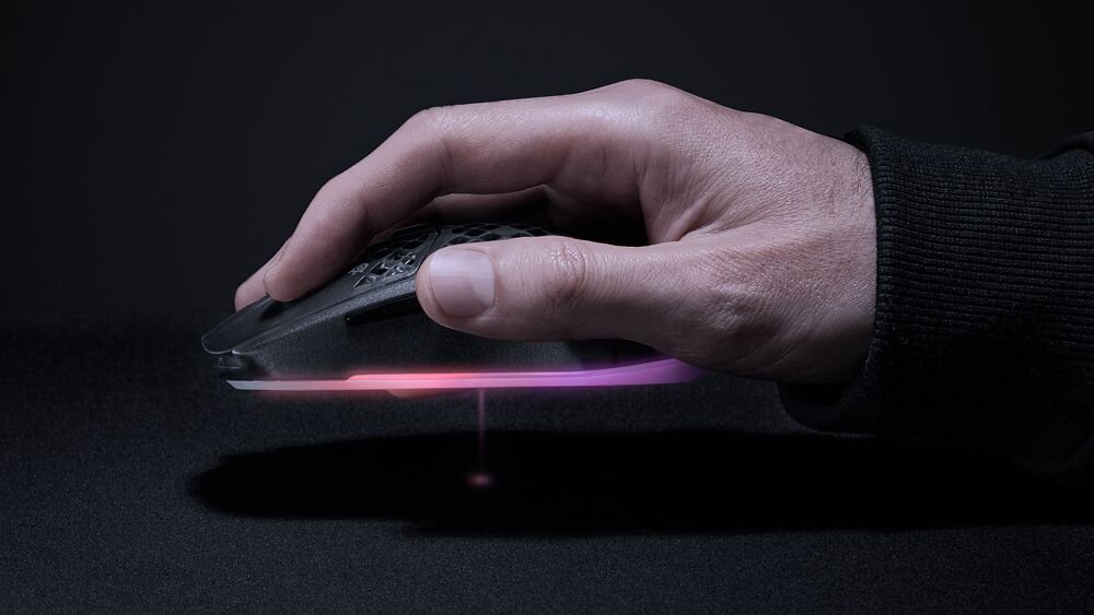 Ręka trzyma mysz komputerową z podświetleniem RGB na ciemnym tle. Mysz ma perforowaną powierzchnię i emituje różnokolorowe światło od spodu.