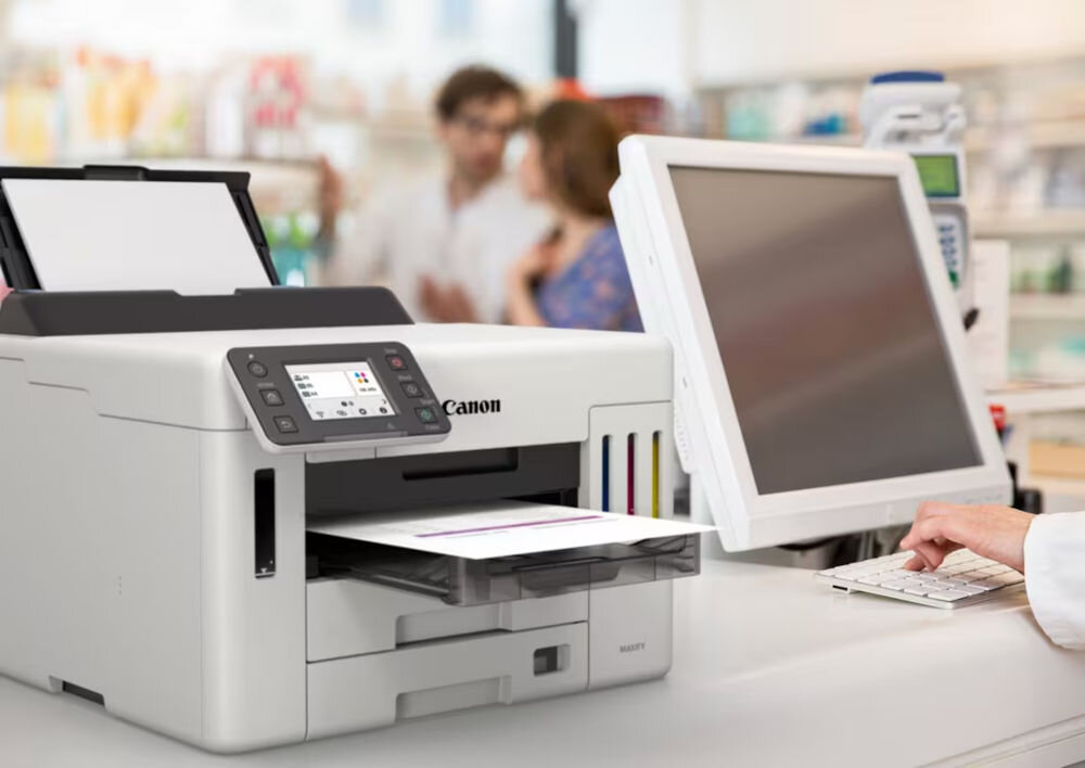 Na obrazie znajduje się urządzenie wielofunkcyjne Canon z wyświetlaczem i otwartą tacą na papier. Obok urządzenia widoczny jest monitor i klawiatura, a w tle rozmyte postacie.