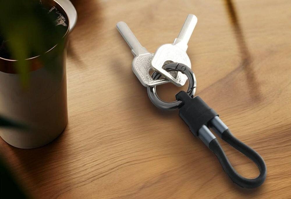 Na drewnianym blacie leży zestaw kluczy z przypiętym czarnym kablem USB-C w formie breloka. Obok znajduje się metalowy kubek z napojem.