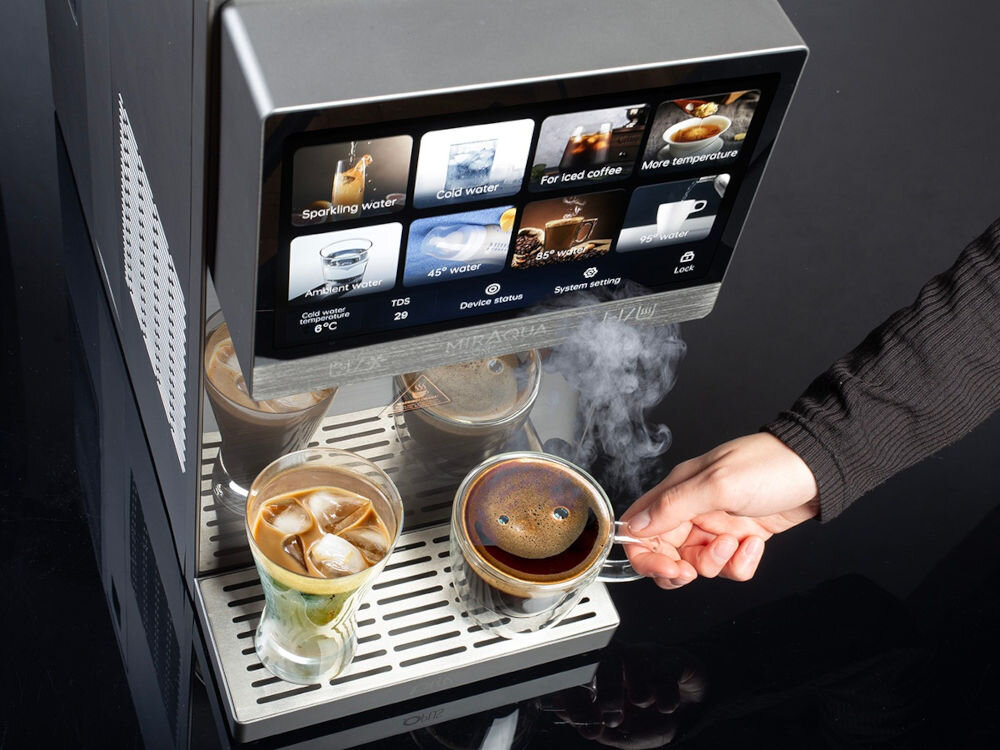 Na obrazie widoczny jest dystrybutor wody z ekranem dotykowym, na którym wyświetlane są różne opcje napojów, takie jak \'Sparkling water\', \'Cold water\', \'For iced coffee\'. Pod urządzeniem znajdują się szklanki z napojami, a jedna z nich jest napełniana gorącą kawą.