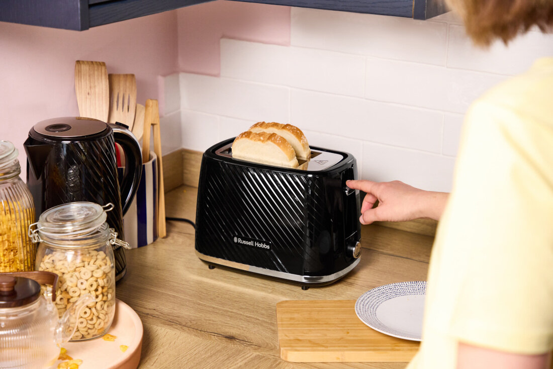 Na blacie kuchennym stoi czarny toster z logo \'Russell Hobbs\', w którym znajduje się kromka chleba. Obok tostera widoczne są słoiki z płatkami śniadaniowymi i czajnik elektryczny.