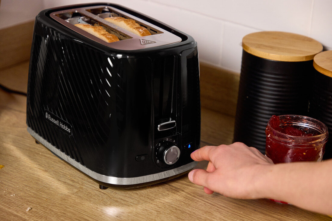 Czarny toster z logo \'Russell Hobbs\' na blacie kuchennym, z dwoma kromkami chleba w środku. Obok stoi słoik z dżemem, a ręka dotyka pokrętła tostera.